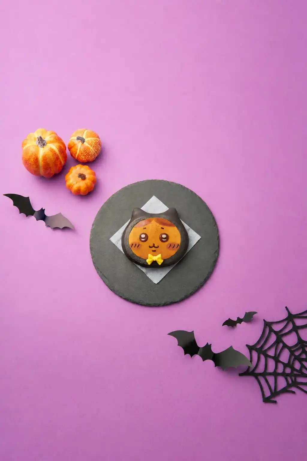 A single Chiikawa Bakery black-cat Halloween bread on a slate plate with mini pumpkins, bat cut-outs and a paper cobweb