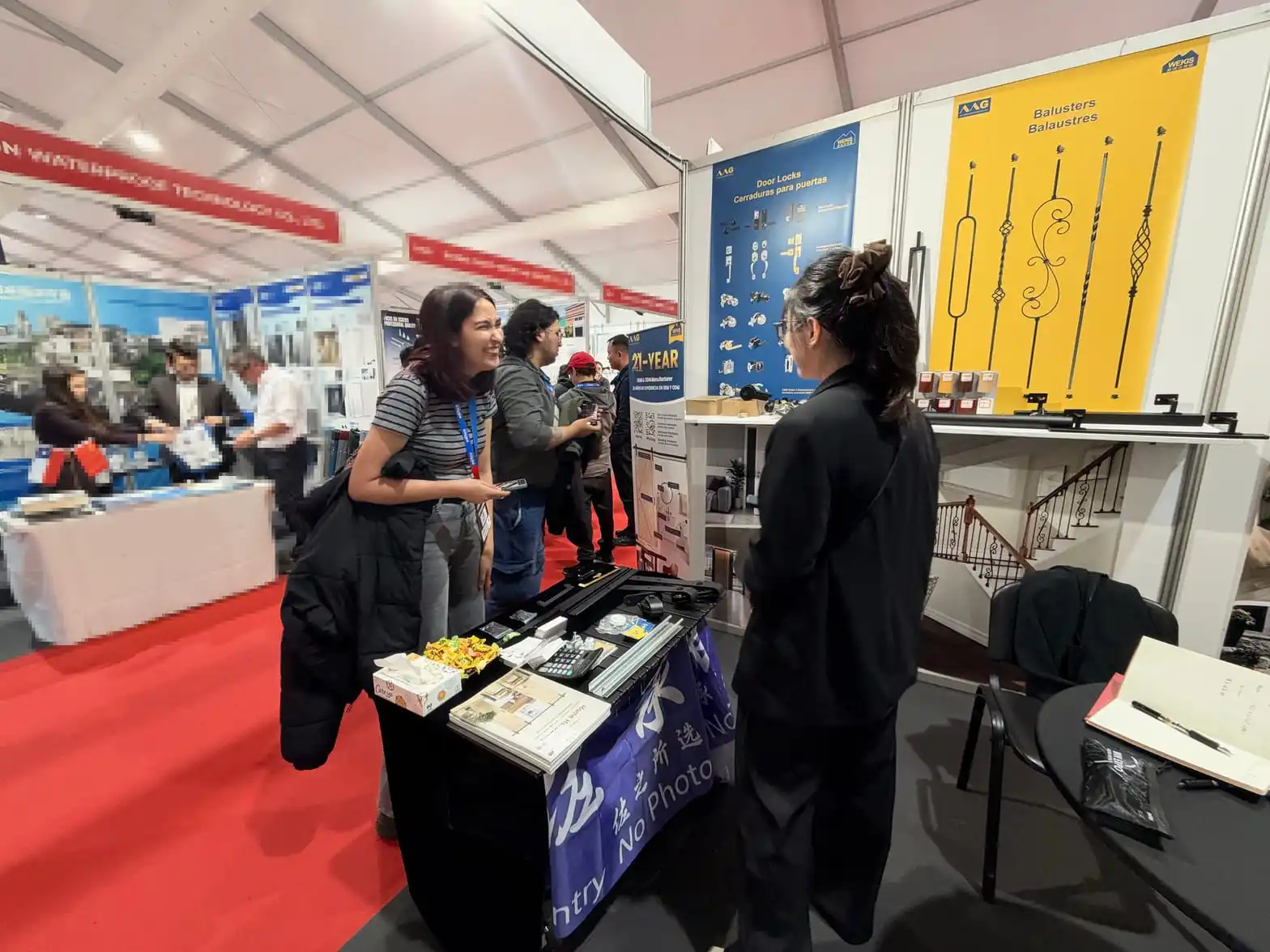 Un membre de l'équipe Wekis discute avec deux visiteurs près du stand présentant les serrures de porte et les balustres à l'EDIFICA 2026.