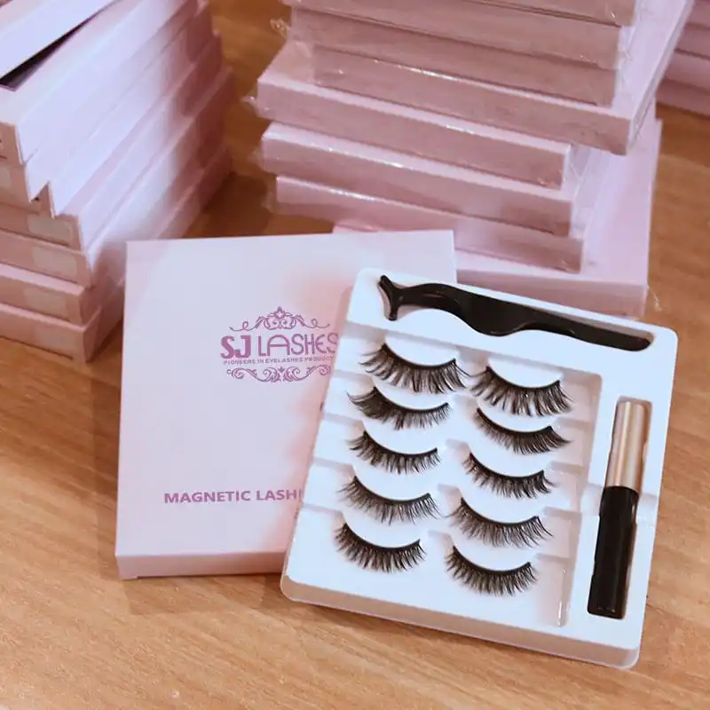 Magnetic lash and eyeliner kit presented on a wooden table with stacked pink boxes in the background, showing the tray contents and the branded box lid - magnetic lash kit presentation