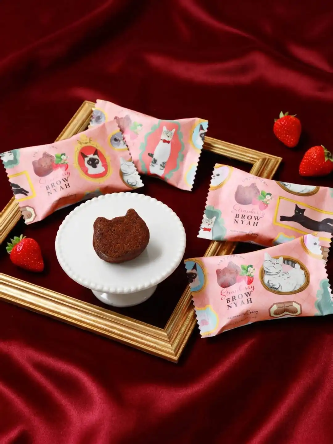 Cat-shaped strawberry brownie on a small cake stand with pink wrappers and strawberries inside a gold frame