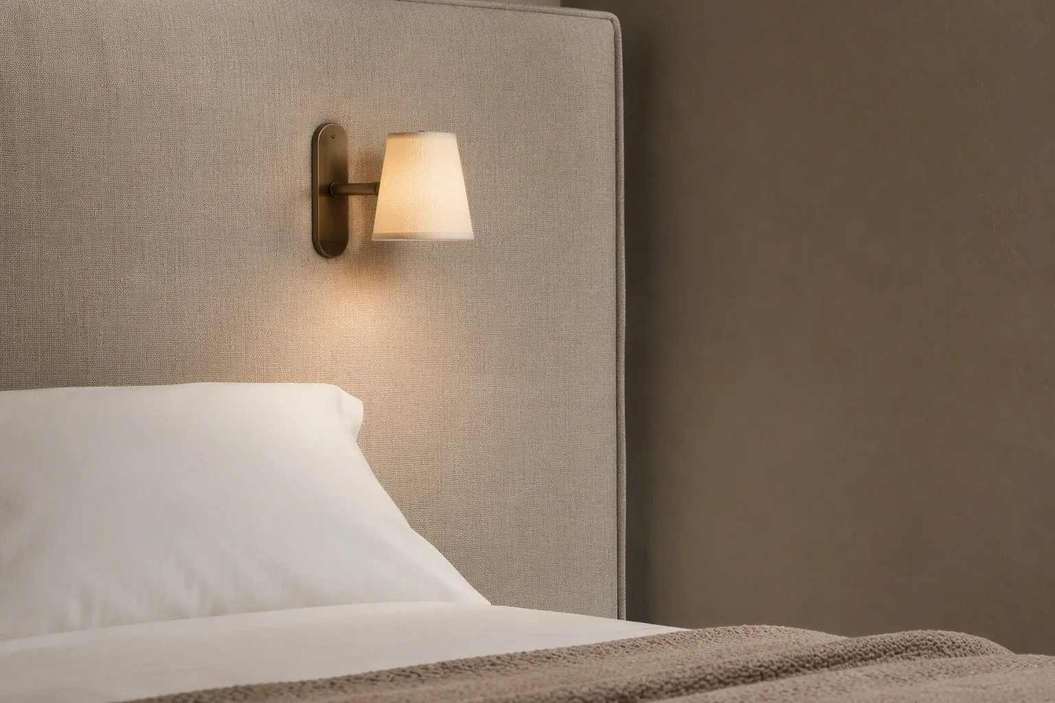 Brass wall-mounted reading light with a linen shade on an upholstered hotel headboard