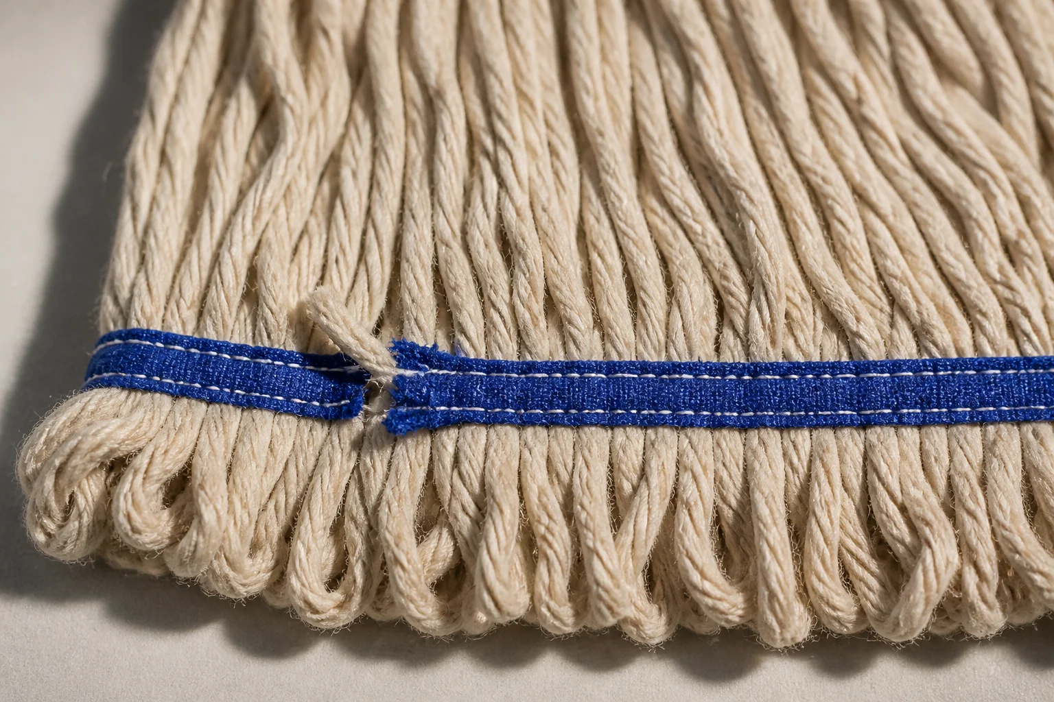 Close view of a blue woven tail band stitched across natural cotton mop yarn, with one section of stitching broken and yarn escaping through the gap