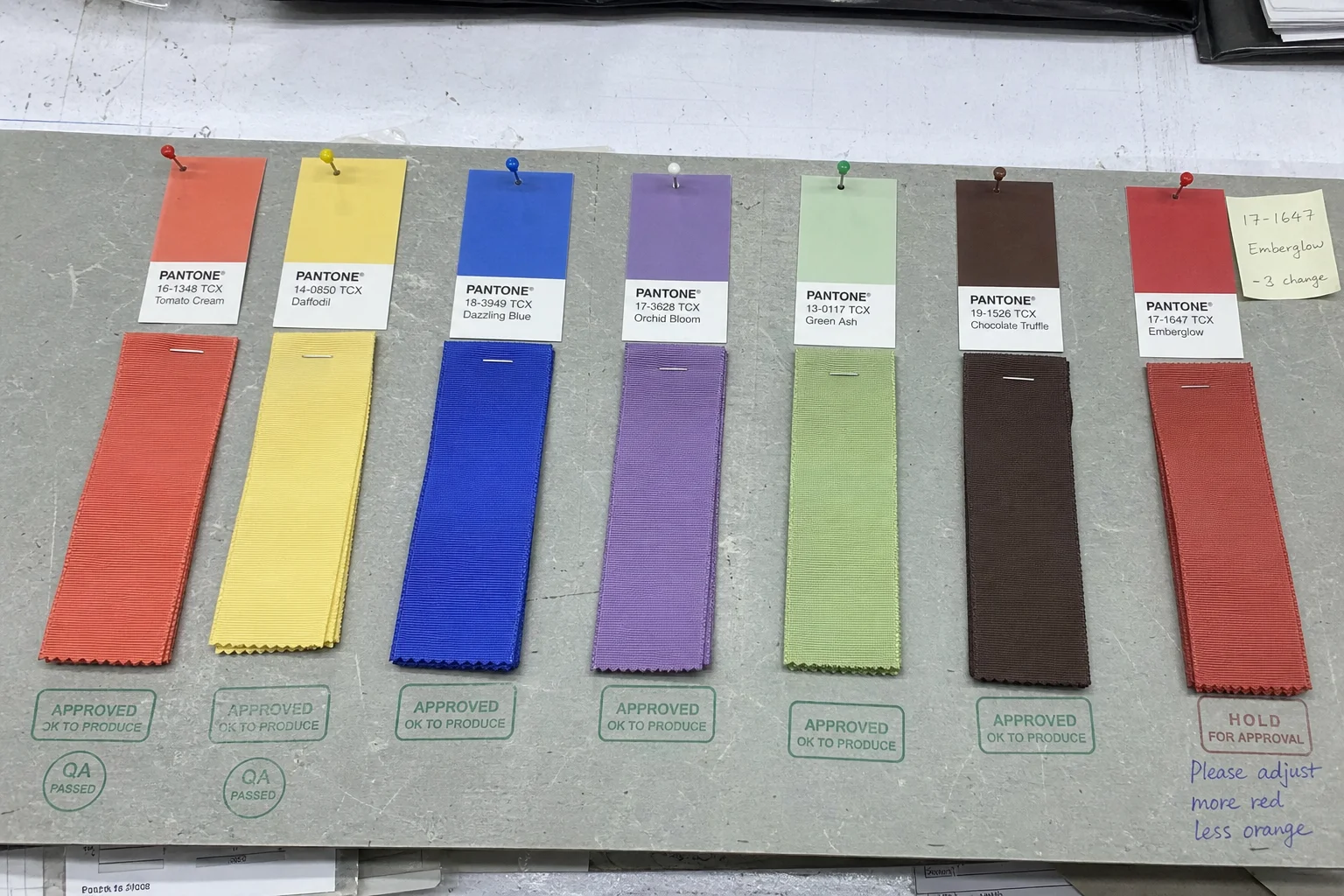 Documentary B2B product photo row colour approval sample ribbons factory quality inspection table each sample 30cm length ribbon new seasonal colour pinned beside Pantone dye reference card showing target some samples green approval stamp one handwritten notes adjustment natural overhead inspection lighting factual colour development documentation