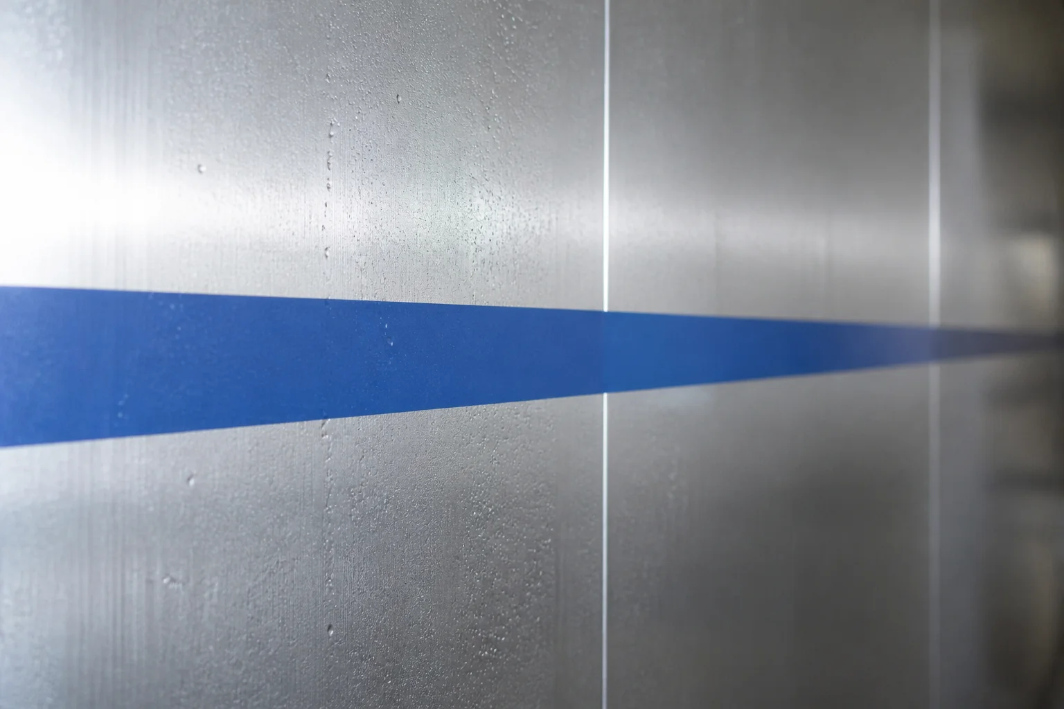 Brushed stainless steel wash-down wall with fine condensation droplets and a single matte blue identification stripe