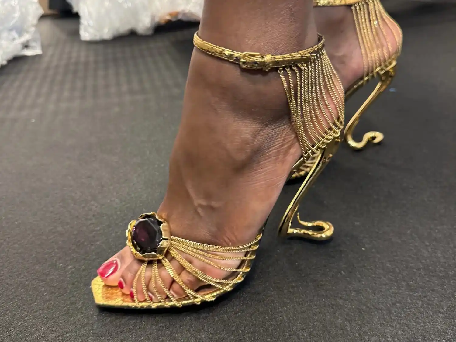 Backstage close-up: gilded chains meeting a large dark purple jewel at the centre of the vamp, on a snake-coil heel