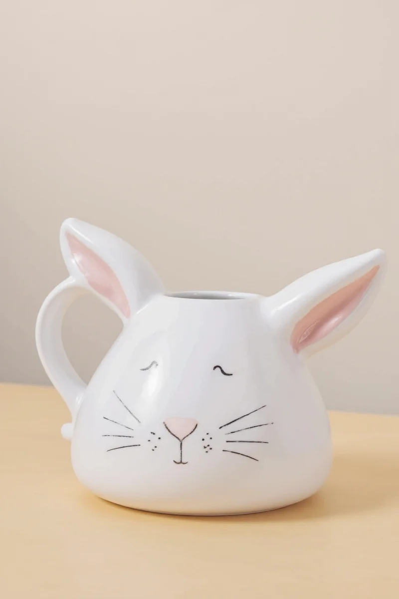 Bunny rabbit pitcher filled with small white daisies and placed next to a woven basket on a rustic table.