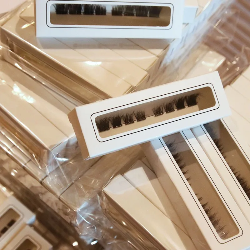 Stacks of white lash boxes with printed rule windows, each showing one row of clusters on a paper tray, packed together on a factory bench - paper tray lash insert