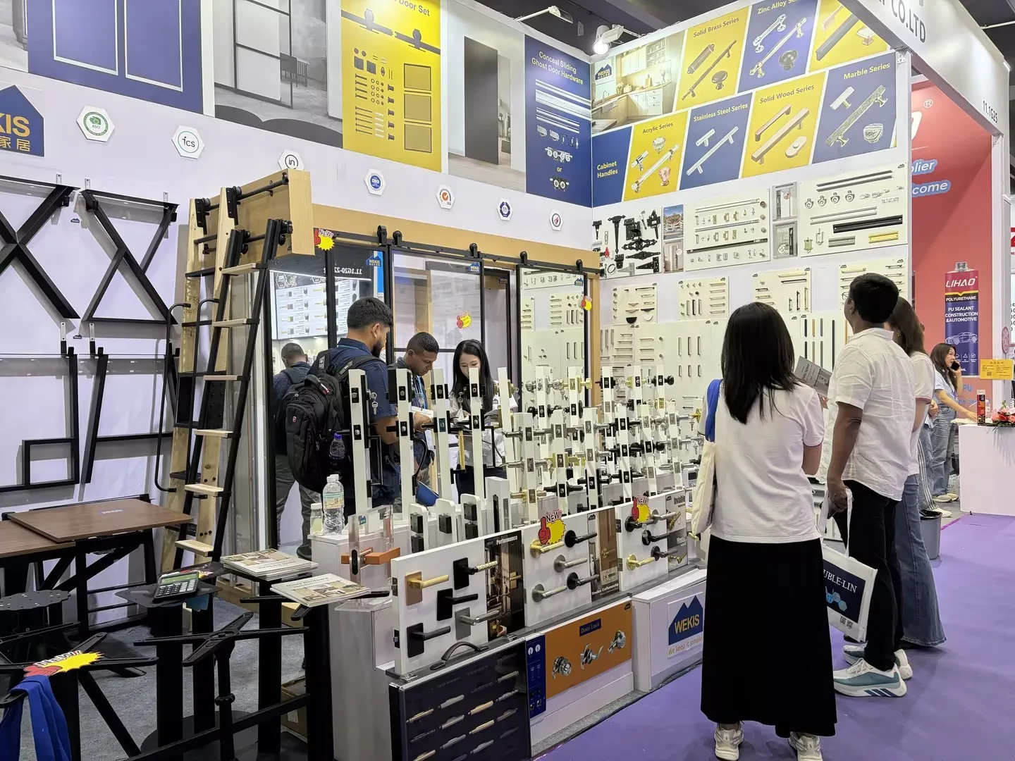 Des acheteurs examinent attentivement le panneau mural de poignées de meubles avec un membre de l'équipe Wekis sur le stand de la Foire de Canton.