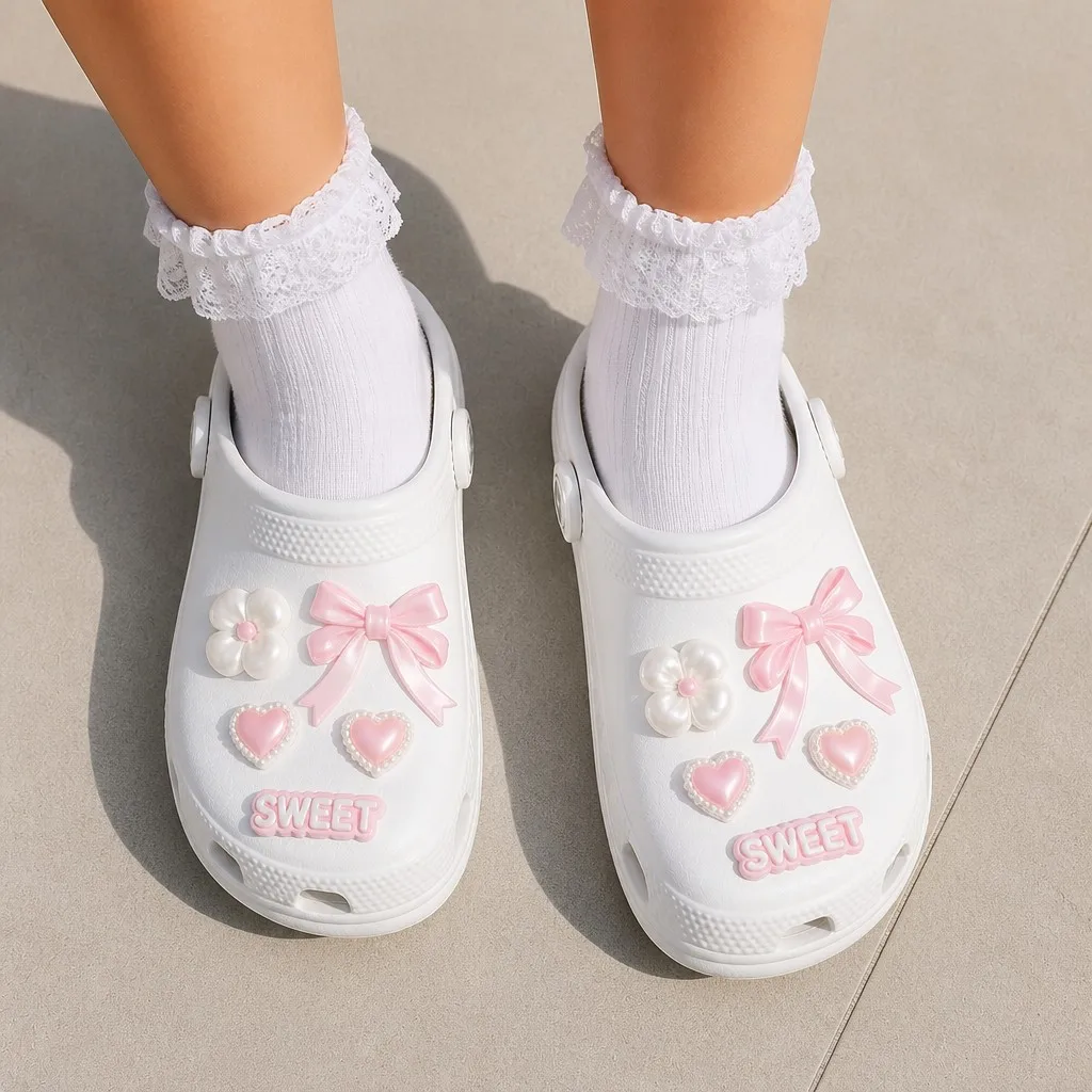 Worn pair of white clogs with bow, pearl flower and heart charms over ruffle-trimmed lace socks