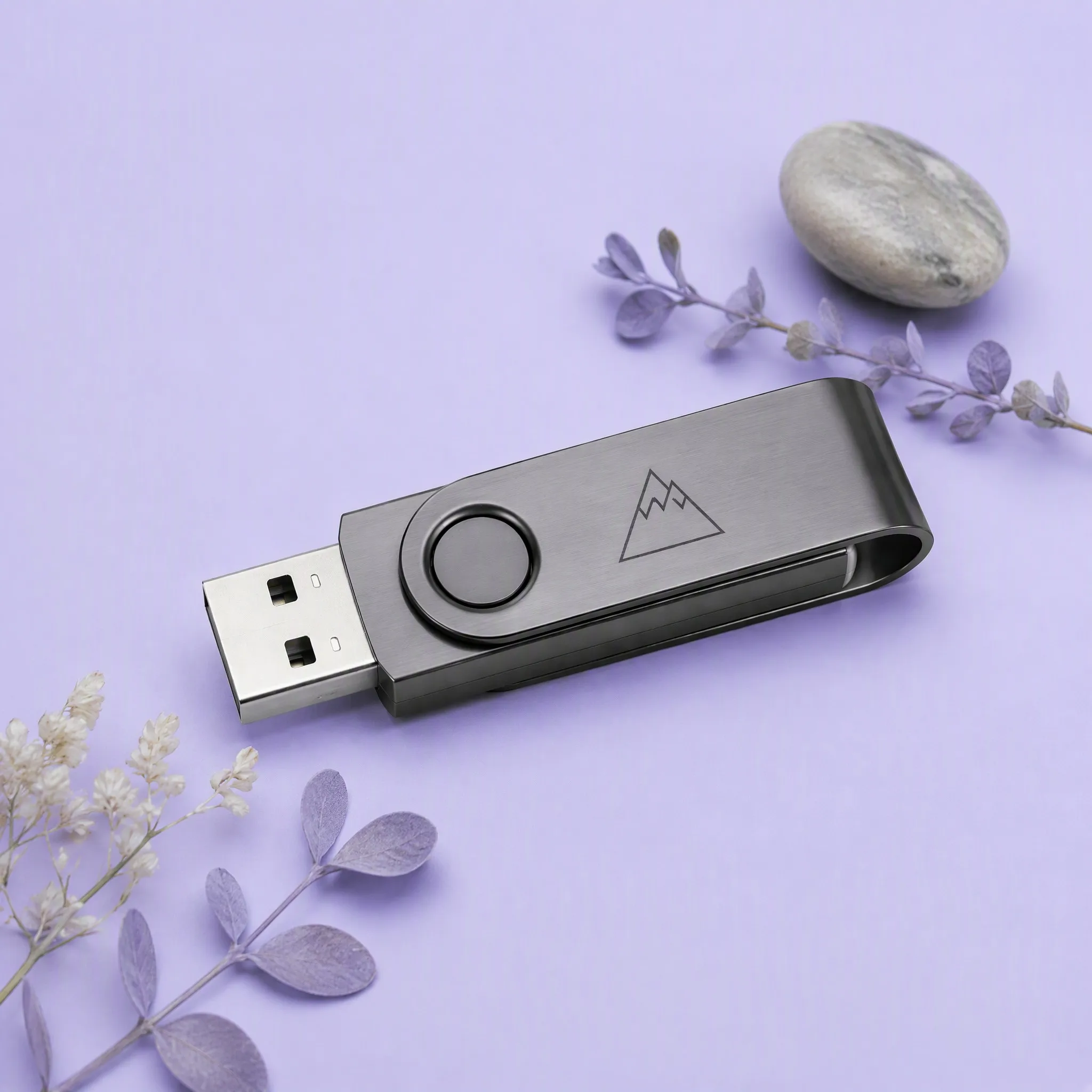 Titanium Gunmetal Grey Swivel USB Flash Drive with Mountain Peak Laser Engraving beside Dried Purple Leaves and River Pebble